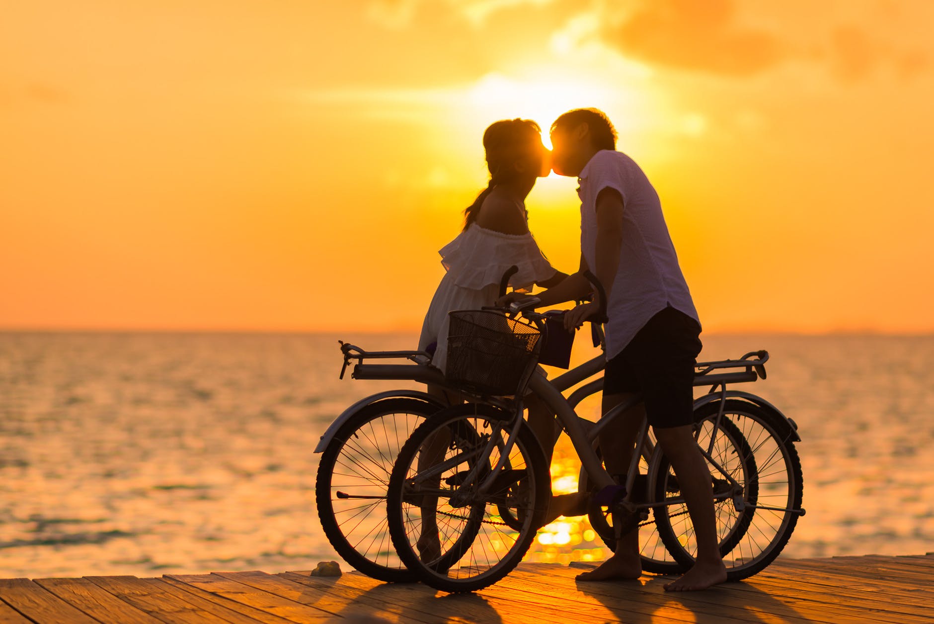 photography of man wearing white t shirt kissing a woman while holding bicycle on river dock during sunset